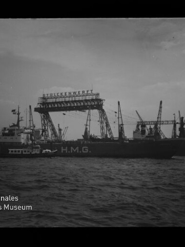 Schwarzweiß-Foto eines Frachtschiffs mit dem Schriftzug H.M.G. vor einer Werft, über der ein großer Gitterkran mit dem Schriftzug „Stülckenwerft“ steht; ein Schlepper an der Seite, Wasser im Vordergrund und das Logo des Internationalen Maritimen Museums Hamburg unten links.
