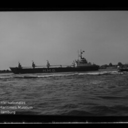 Schwarzweißaufnahme eines Frachtschiffs mit der Aufschrift H.M.G. auf der Bordwand, das auf leicht kabbeliger See an einer Kaimauer vorbeifährt; links unten sichtbar das Logo und die Beschriftung des Internationalen Maritimen Museums Hamburg.