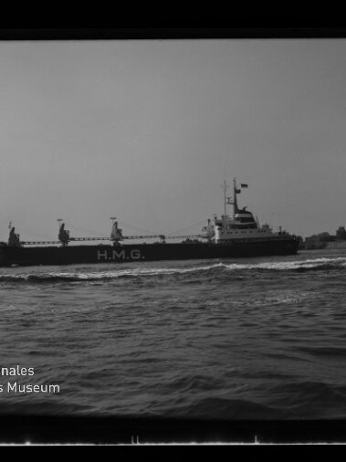 Schwarzweißaufnahme eines Frachtschiffs mit der Aufschrift H.M.G. auf der Bordwand, das auf leicht kabbeliger See an einer Kaimauer vorbeifährt; links unten sichtbar das Logo und die Beschriftung des Internationalen Maritimen Museums Hamburg.