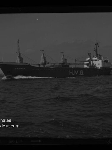 Schwarzweiß-Foto eines Frachtschiffs mit großer Aufschrift „H.M.G.“ am Rumpf und mehreren Ladekränen, das mit Bugwelle über die See fährt; im Hintergrund flacher Küstenstreifen und ein kleines Segelboot, links unten Museumswasserzeichen.