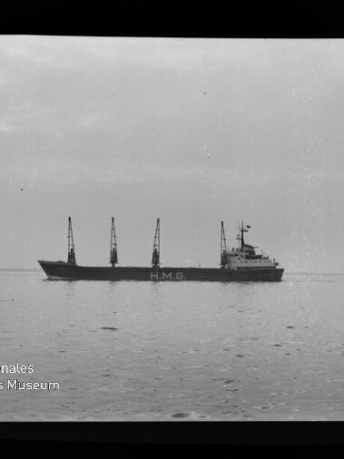 Schwarzweißaufnahme eines Frachtschiffs mit der Aufschrift H.M.G. und mehreren Ladekränen, das ruhig über spiegelglattes Wasser vor einem flachen Horizont fährt.
