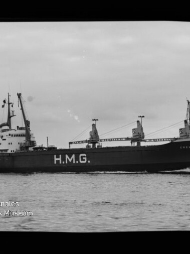 Schwarzweißfoto eines Frachtschiffs mit großer Aufschrift H.M.G. am Rumpf, drei Ladekräne und das Deckshaus achtern sichtbar, das auf ruhiger See fährt; links unten Wasserzeichen des Internationalen Maritimen Museums Hamburg.