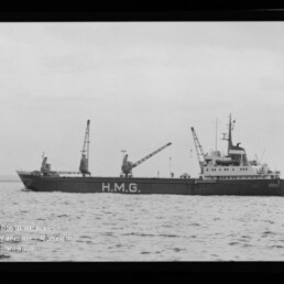Schwarzweiß-Foto eines Frachtschiffs mit großer Aufschrift H.M.G. am Rumpf, drei Ladekräne auf dem Deck und Aufbau am Heck, das auf ruhiger See fährt; schwarzer Bildrand und Museumswasserzeichen unten links sind sichtbar.