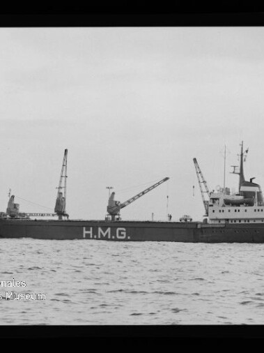 Schwarzweiß-Foto eines Frachtschiffs mit großer Aufschrift H.M.G. am Rumpf, drei Ladekräne auf dem Deck und Aufbau am Heck, das auf ruhiger See fährt; schwarzer Bildrand und Museumswasserzeichen unten links sind sichtbar.