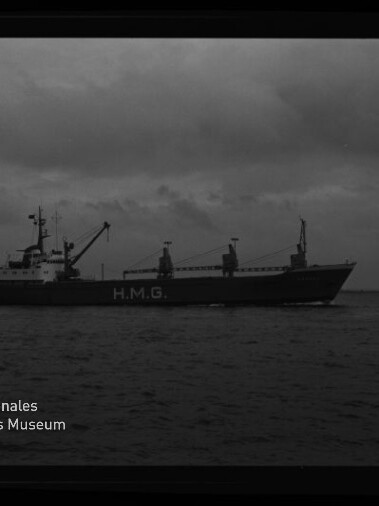 Schwarzweiß-Foto eines Frachtschiffs mit der Aufschrift H.M.G. auf dem Rumpf, das bei bedecktem Himmel auf ruhiger See vorbeifährt; links unten das Logo des Internationalen Maritimen Museums Hamburg und am rechten Bildrand der Filmrand mit der Aufschrift AGFA LISS sichtbar.