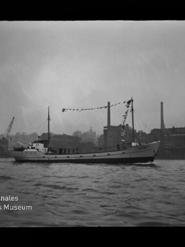 Schwarzweißaufnahme eines geschmückten Küsten- oder Arbeitsschiffs mit Flaggenbändern, das auf ruhigem Wasser vor einer industriellen Hafenfront mit Kränen und Fabrikschornsteinen fährt; Logowasserzeichen des Internationalen Maritimen Museums Hamburg in der linken unteren Ecke.