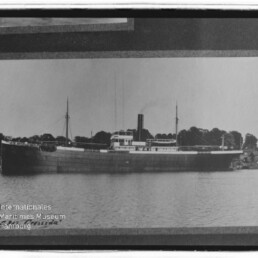 Schwarzweiß-Foto eines historischen Dampffrachters mit mittigem Schornstein und zwei Masten, am Ufer vor einer Baumreihe liegend und ruhiger Wasserfläche im Vordergrund, mit Museum-Logo unten links.