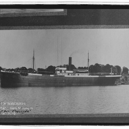 Schwarzweiß-Foto eines historischen Dampffrachters mit mittigem Schornstein und zwei Masten, am Ufer vor einer Baumreihe liegend und ruhiger Wasserfläche im Vordergrund, mit Museum-Logo unten links.