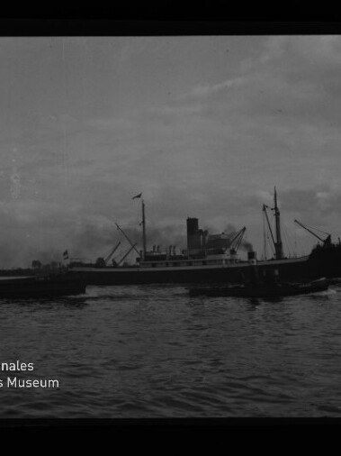 Schwarzweiß-Foto eines größeren Frachtdampfers mit markantem Schornstein und Ladekränen, neben einem kleineren Begleitboot auf ruhiger See unter bewölktem Himmel; unten links das Logo und Schriftzug des Internationalen Maritimen Museums Hamburg.