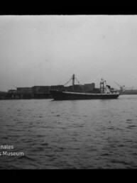 Schwarzweißaufnahme eines Frachtschiffs, das auf ruhigem Wasser vor einer Hafenbebauung mit Kränen und Lagerhäusern fährt.