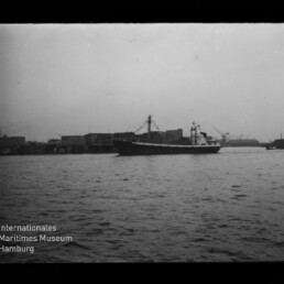 Schwarzweißaufnahme eines Frachtschiffs, das auf ruhigem Wasser vor einer Hafenbebauung mit Kränen und Lagerhäusern fährt.