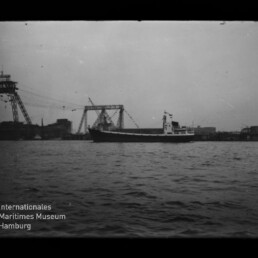Schwarzweißaufnahme eines Frachtschiffs auf ruhigem Wasser vor einem Industriehafen mit großen Auslegerkränen und Hafenanlagen am Horizont.