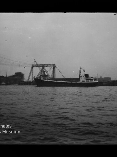 Schwarzweißaufnahme eines Frachtschiffs auf ruhigem Wasser vor einem Industriehafen mit großen Auslegerkränen und Hafenanlagen am Horizont.
