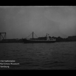 Schwarzweißaufnahme eines Frachtschiffs auf dem Wasser vor einer Industrie-Küste mit stählerner Brückenkonstruktion links und dunklem Himmel; unten links das Logo des Internationalen Maritimen Museums Hamburg.