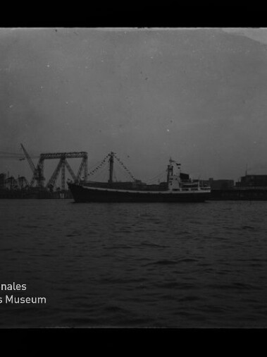 Frachtschiff auf ruhigem Wasser vor der Silhouette eines Industriehafens mit Kränen und einer großen Hubbrücke, dunkle kontrastarme Aufnahme mit Museumslogo unten links.