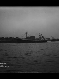 Schwarzweißaufnahme eines Frachtschiffs mit beflaggtem Mast, das in einem Hafenbecken vor einer Kaimauer mit Lagerhäusern, Kränen und einem Fahrwasserturm liegt; im Vordergrund welliges Wasser und unten links das Logo des Internationalen Maritimen Museums Hamburg.