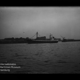 Schwarzweißaufnahme eines Frachtschiffs mit beflaggtem Mast, das in einem Hafenbecken vor einer Kaimauer mit Lagerhäusern, Kränen und einem Fahrwasserturm liegt; im Vordergrund welliges Wasser und unten links das Logo des Internationalen Maritimen Museums Hamburg.