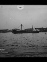 Schwarzweiß-Foto eines Frachtschiffs mit geschmückten Signalflaggen, seitlich auf der Wasseroberfläche vor einem Kai mit Industrieanlagen und einer Hubbrücke; unten links das Wasserzeichen des Internationalen Maritimen Museums Hamburg.