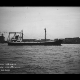 Schwarzweiß-Foto eines Frachtschiffs mit Aufbauten mittschiffs und Mast, das auf bewegter See an einem Industriehafen mit Ladekränen im Hintergrund entlangfährt; unten links sichtbar das Logo des Internationalen Maritimen Museums Hamburg.