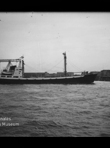 Schwarzweiß-Foto eines Frachtschiffs mit Aufbauten mittschiffs und Mast, das auf bewegter See an einem Industriehafen mit Ladekränen im Hintergrund entlangfährt; unten links sichtbar das Logo des Internationalen Maritimen Museums Hamburg.