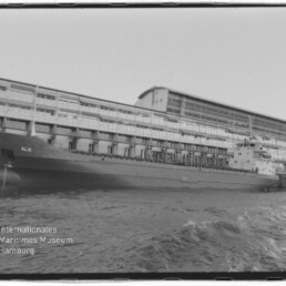 Schwarzweißaufnahme eines langen Frachtschiffs mit der Aufschrift ALK, längsseits an einem mehrstöckigen Industrie-Kai festgemacht; im Vordergrund bewegtes Wasser und unten links das Wasserzeichen des Internationalen Maritimen Museums Hamburg.