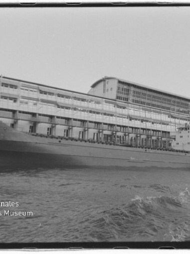 Schwarzweißaufnahme eines langen Frachtschiffs mit der Aufschrift ALK, längsseits an einem mehrstöckigen Industrie-Kai festgemacht; im Vordergrund bewegtes Wasser und unten links das Wasserzeichen des Internationalen Maritimen Museums Hamburg.