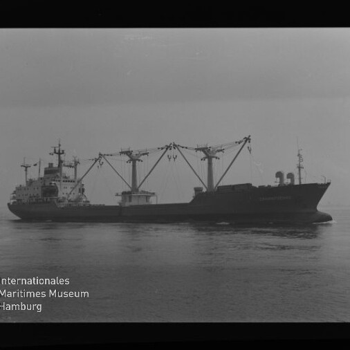Schwarzweiß-Seitenansicht eines Frachtschiffs mit mehreren markanten Ladekränen über dem Laderaum, das ruhig auf dem Meer fährt; in der linken unteren Ecke Logo und Schriftzug des Internationalen Maritimen Museums Hamburg sichtbar.