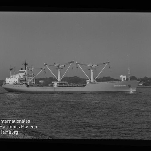 Schwarzweiß-Bild eines Frachtschiffs mit drei großen Ladekränen, das vor einer flachen Küstenlinie fährt; links unten sichtbares Logo und Schriftzug des Internationalen Maritimen Museums Hamburg sowie deutlicher Filmrand mit Markierungen.