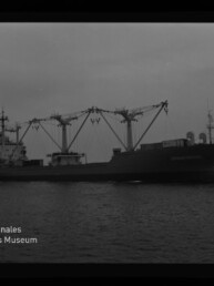 Seitenansicht eines Frachtschiffs auf ruhiger See mit mehreren markanten Ladekränen und Aufbauten, schwarzweiß aufgenommen; Logo des Internationalen Maritimen Museums Hamburg unten links.