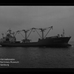 Seitenansicht eines Frachtschiffs auf ruhiger See mit mehreren markanten Ladekränen und Aufbauten, schwarzweiß aufgenommen; Logo des Internationalen Maritimen Museums Hamburg unten links.
