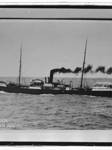 Schwarzweiß-Foto eines seitlich fahrenden Dampfschiffs mit mittigem Schornstein, der schwarzen Rauch ausstößt, zwei Masten, Flagge am Heck und leicht bewegter See, mit weißem Bildrand und Museumslogo unten links.