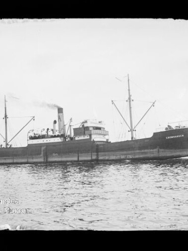 Seitenansicht eines dampfbetriebenen Frachtschiffs mit einem einzelnen Schornstein, zwei Masten und Ladegeschirr, das ruhig auf dem Wasser liegt; unten links sichtbares Museumswasserzeichen.