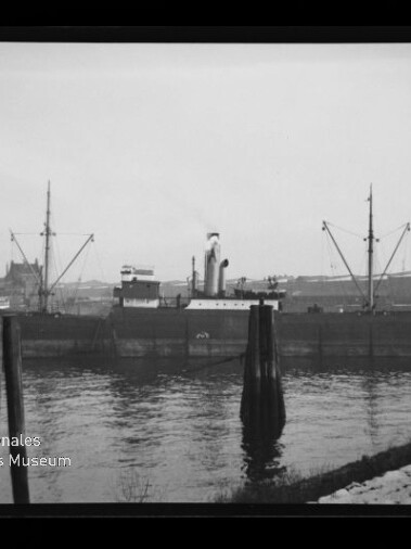 Schwarzweißaufnahme eines Frachtschiffs mit Schornstein und Ladebäumen an einem Industriehafen, im Vordergrund Holzdalben im Wasser und links unten das Logo des Internationalen Maritimen Museums Hamburg.