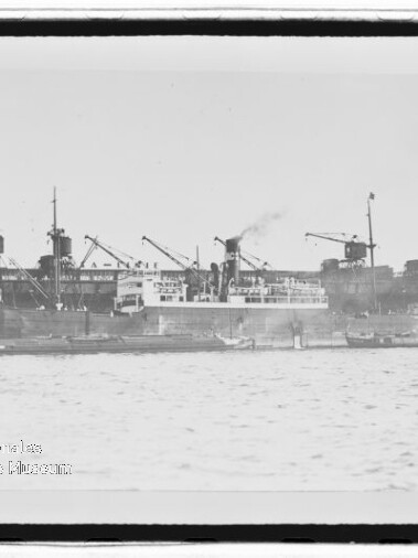 Schwarzweiß-Aufnahme eines Dampfschiffs, das an einem Industriequai mit mehreren Ladekränen liegt; Rauch steigt aus dem Schornstein und kleine Boote fahren im Vordergrund auf dem Wasser.