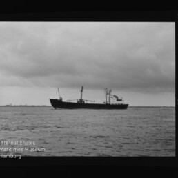 Schwarzweiß-Foto eines Frachtschiffs in Seitenansicht auf ruhiger See unter wolkenverhangenem Himmel, mit Filmrand und Wasserzeichen ‚Internationales Maritimes Museum Hamburg‘ links unten.