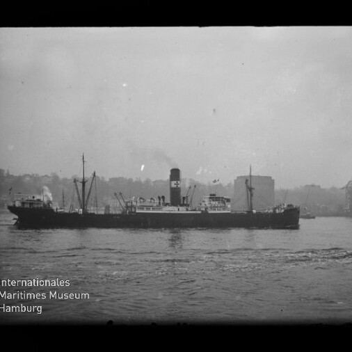 Schwarzweißfoto eines längs fahrenden Dampffrachters mit hohem Schornstein und Rauchsäule, vor einer Hafenansicht mit Kaikränen und Gebäuden; Wasser im Vordergrund und Museums-Logo unten links.