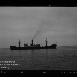 Schwarzweißaufnahme eines dampfbetriebenen Frachtschiffs in Seitenansicht auf ruhiger See, dunkle Silhouette mit rauchendem Schornstein, flache Küstenlinie am Horizont sowie sichtbare Filmrandmarkierungen und Wasserzeichen des Internationalen Maritimen Museums Hamburg