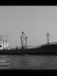 Schwarz-weiße Seitenansicht eines großen Frachtschiffs auf ruhiger See mit mehreren Ladekränen und Aufbau am Heck; unten links das Wasserzeichen des Internationalen Maritimen Museums Hamburg.