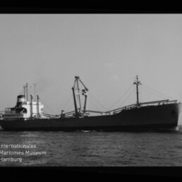 Schwarz-weiße Seitenansicht eines großen Frachtschiffs auf ruhiger See mit mehreren Ladekränen und Aufbau am Heck; unten links das Wasserzeichen des Internationalen Maritimen Museums Hamburg.