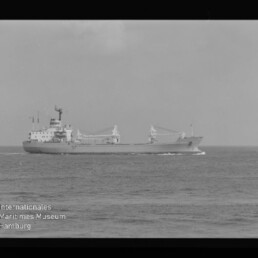 Schwarzweißaufnahme eines Frachtschiffs in Fahrt auf ruhiger See, seitlich zu sehen mit zwei Ladekränen und mittig liegenden Aufbauten vor dem Horizont; links unten ist ein Wasserzeichen des Internationalen Maritimen Museums Hamburg und ein schwarzer Filmrand erkennbar.
