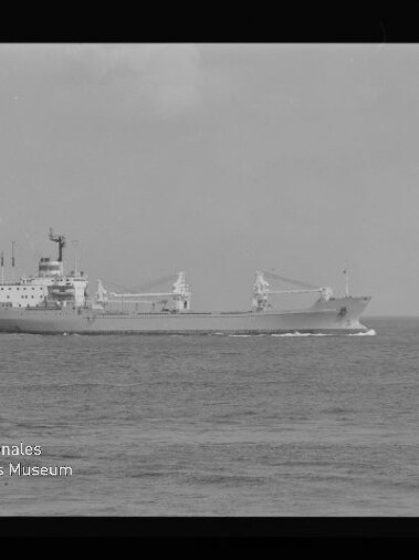 Schwarzweißaufnahme eines Frachtschiffs in Fahrt auf ruhiger See, seitlich zu sehen mit zwei Ladekränen und mittig liegenden Aufbauten vor dem Horizont; links unten ist ein Wasserzeichen des Internationalen Maritimen Museums Hamburg und ein schwarzer Filmrand erkennbar.