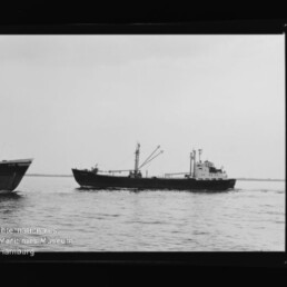 Schwarzweiß-Foto eines Frachtschiffs mit Ladekränen und achternliegenden Aufbauten, das auf ruhiger See fährt; links am Bildrand der Bug eines weiteren Schiffs sowie Filmrand und Wasserzeichen des Internationalen Maritimen Museums Hamburg sichtbar.