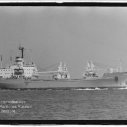 Schwarzweiß-Foto eines Frachtschiffs in Seitenansicht mit Aufbauten achtern und mehreren Deckskränen, das nach rechts fahrend auf ruhiger See eine Bugwelle zieht; am unteren linken Bildrand Wasserzeichen des Internationalen Maritimen Museums Hamburg.