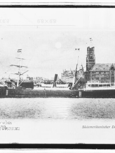 Schwarzweiß-Aufnahme eines südamerikanischen Dampfers namens „Curityba“ mit zwei Masten und Schornstein, liegend an einem Kai vor einem großen historischen Backsteingebäude mit Turm und spitzer Kirchturm-Silhouette, links unten ein rundes Wasserzeichen.