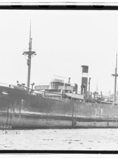 Schwarzweißes Foto eines großen Dampffrachters mit hohem Schornstein und mehreren Masten, seitlich im Hafen festgemacht, Wasser im Vordergrund und Gebäude im Hintergrund, unten links Wasserzeichen des Internationalen Maritimen Museums Hamburg.