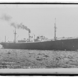 Schwarz-weiß-Seitenaufnahme eines frühen Frachtschiffs mit rauchendem Schornstein und zwei Masten auf leicht welliger See, unten links sichtbar das Wasserzeichen des Internationalen Maritimen Museums Hamburg.