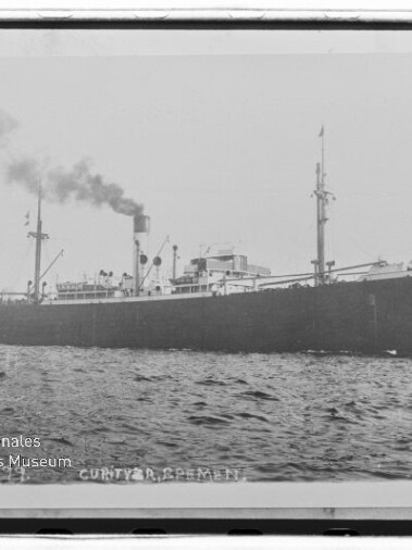 Schwarz-weiß-Seitenaufnahme eines frühen Frachtschiffs mit rauchendem Schornstein und zwei Masten auf leicht welliger See, unten links sichtbar das Wasserzeichen des Internationalen Maritimen Museums Hamburg.