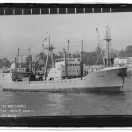 Schwarzweiß-Foto eines Frachtschiffs mit der Aufschrift CUBA, mehreren Masten und Ladekränen, fahrend vor einer Hafenbebauung; Rand des Filmnegativs und Wasserzeichen des Internationalen Maritimen Museums Hamburg sichtbar.