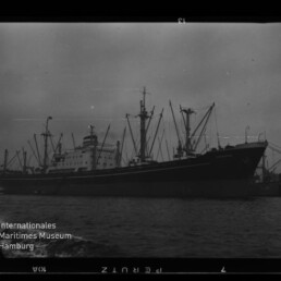 Schwarzweißaufnahme eines großen Frachtschiffs mit mehreren Masten und Ladekränen, am Kai liegend mit Wasser im Vordergrund und dem Wasserzeichen des Internationalen Maritimen Museums Hamburg links unten.