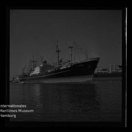 Schwarzweiß-Aufnahme eines großen Frachtschiffs mit Masten und markanter Bugpartie am Kai, ruhige Wasseroberfläche mit Spiegelung; unten links Logo und Schriftzug des Internationalen Maritimen Museums Hamburg.